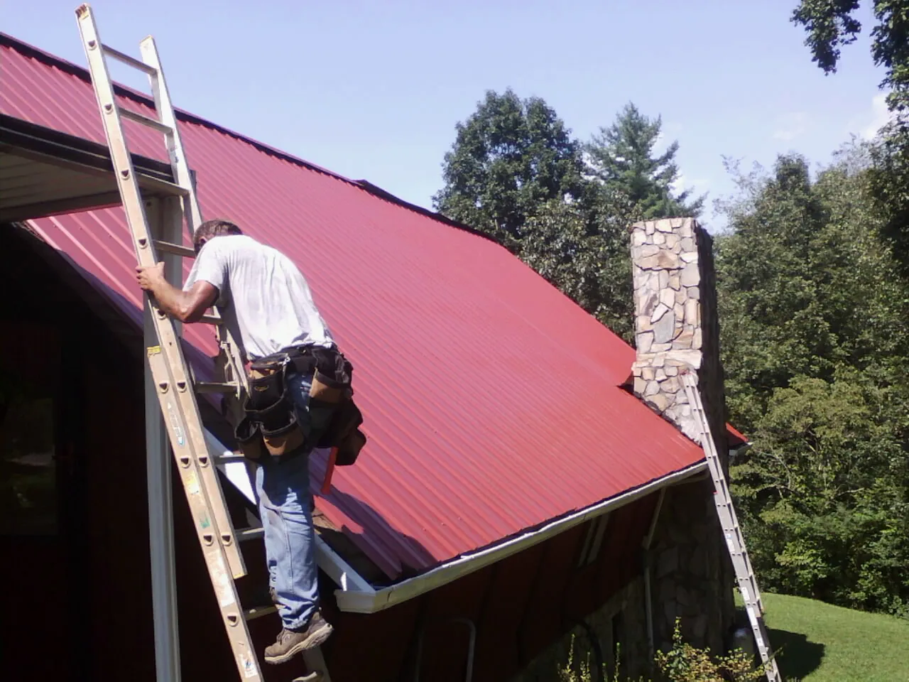 Roofer on a ladder beside a red metal roof and stone chimney — Soffit & Fascia Repair by Roofing Brothers in Rockford